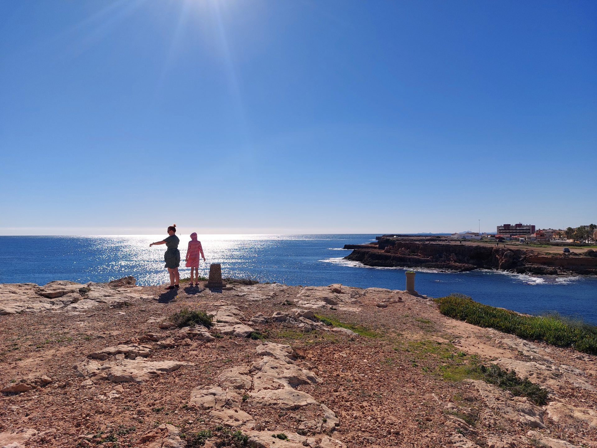 Eine Mutter und ihre Tochter blicken von einem felsigen Strandabschnitt aufs Meer. Sie stehen im Lichtkegel der Sonne, der auf dem Wasser schimmert.